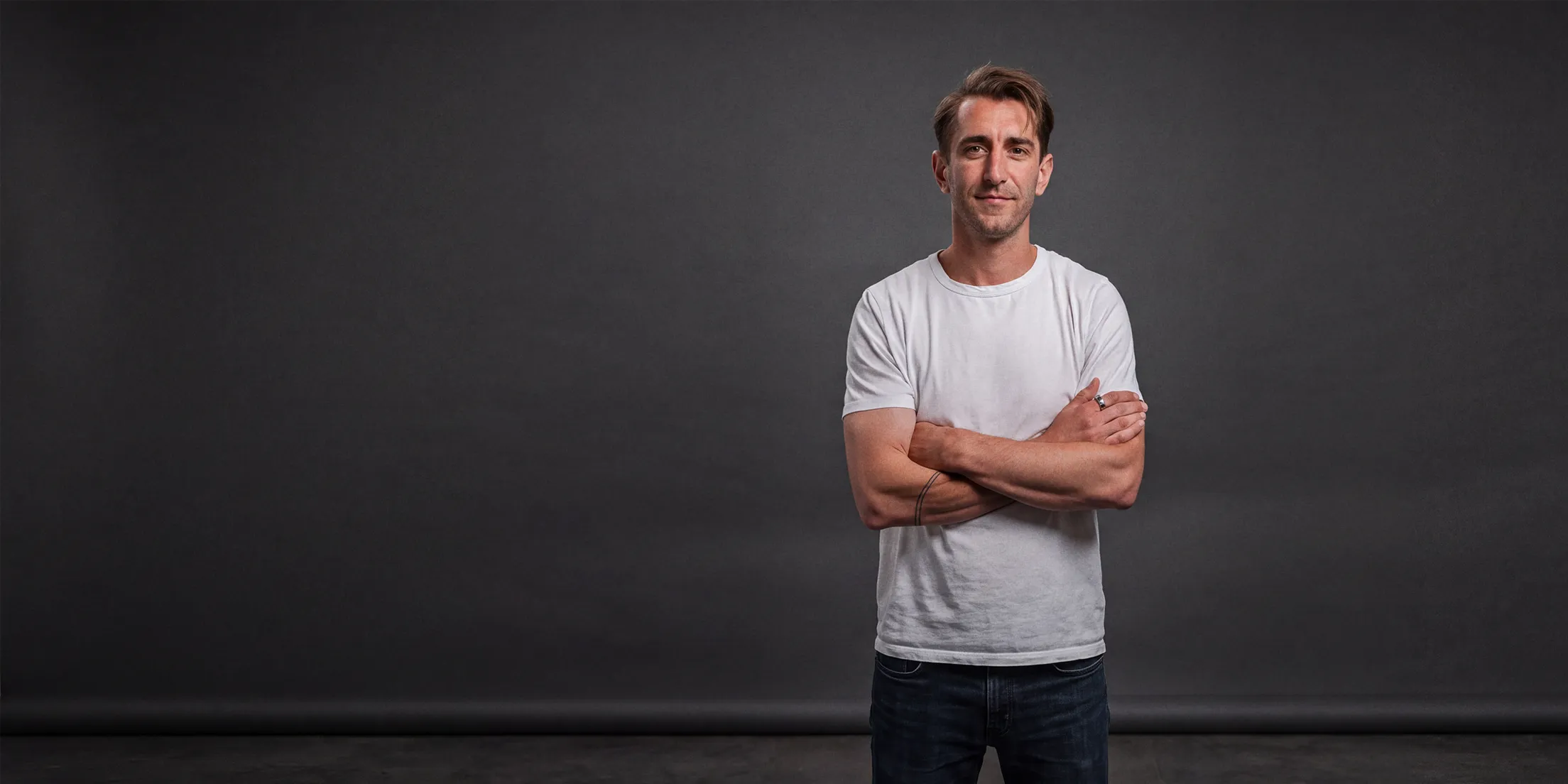 Joel Martin standing with arms crossed in a white t-shirt against a dark grey studio backdrop