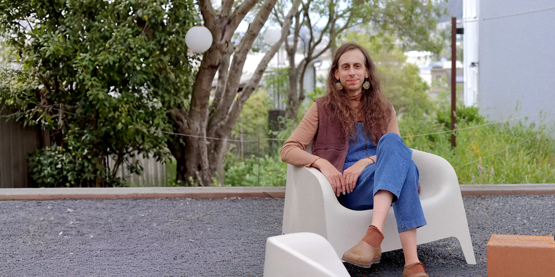 Lily Bodnar sitting in a white molded chair on a gravel patio, trees and a house in the background